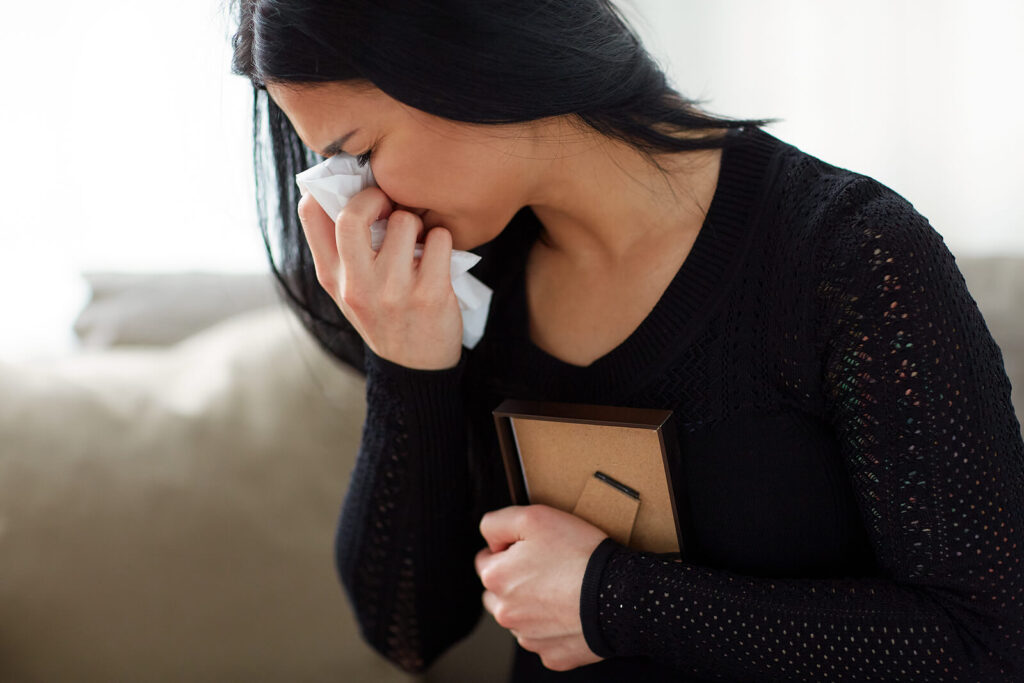 Image of a crying woman clutching a picture frame to her chest. If your grief symptoms are becoming too overwhelming work with a skilled therapist in life transition therapy in Hermosa Beach, CA can help you cope.