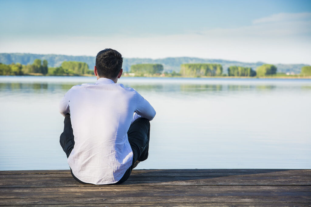 Image of a man sitting by himself on a dock looking out at a lake. Begin coping with your anxiety and find holistic approaches to anxiety with the help of an anxiety therapist in Manhattan Beach, CA.