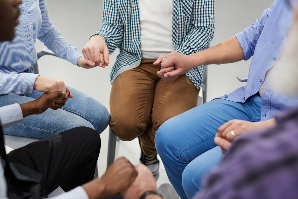 Image of a group of people sitting in a circle holding hands for support. If you find yourself struggling from anxiety, learn how holistic anxiety therapy in Manhattan Beach, CA can help you begin healing.