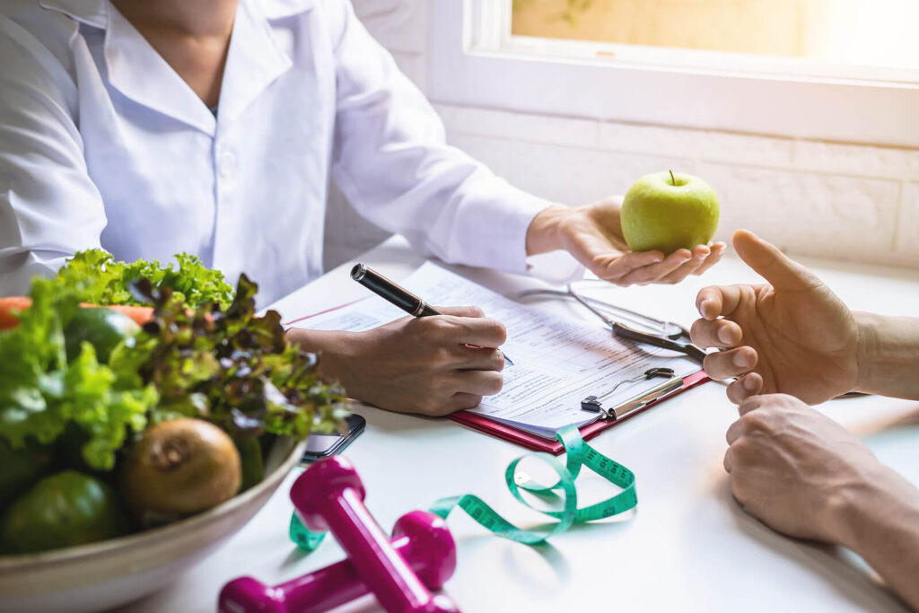 Image of a woman holding an apple and writing while speaking to a man. To overcome your anxiety, you can change your lifestyle and environmental factors. For more support reach out to an anxiety therapist in Manhattan Beach, CA.