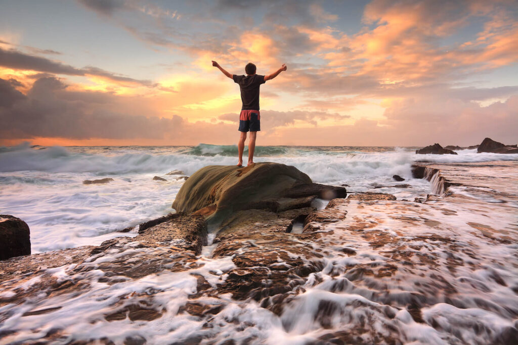 Image of a man standing on a large boulder beside the ocean with his arms raised in the air. This image represents how you can begin breaking free and find healing healing for your anxiety with the help of anxiety therapy in Manhattan Beach, CA.