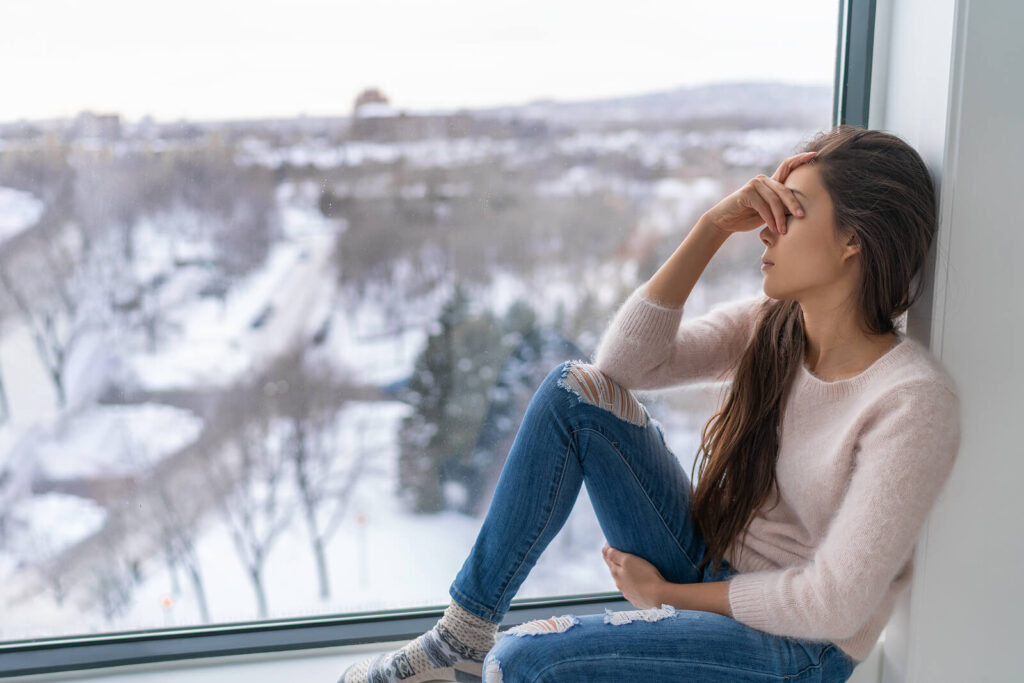 Image of an anxious woman sitting in a window sill covering her face. Find support in healing your anxiety with the help of anxiety therapy in Manhattan Beach, CA. Find holistic healing approaches for anxiety soon.